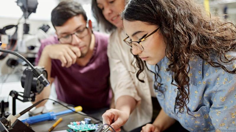 a faculty member (middle) helps a student with an electronics project while another observes
