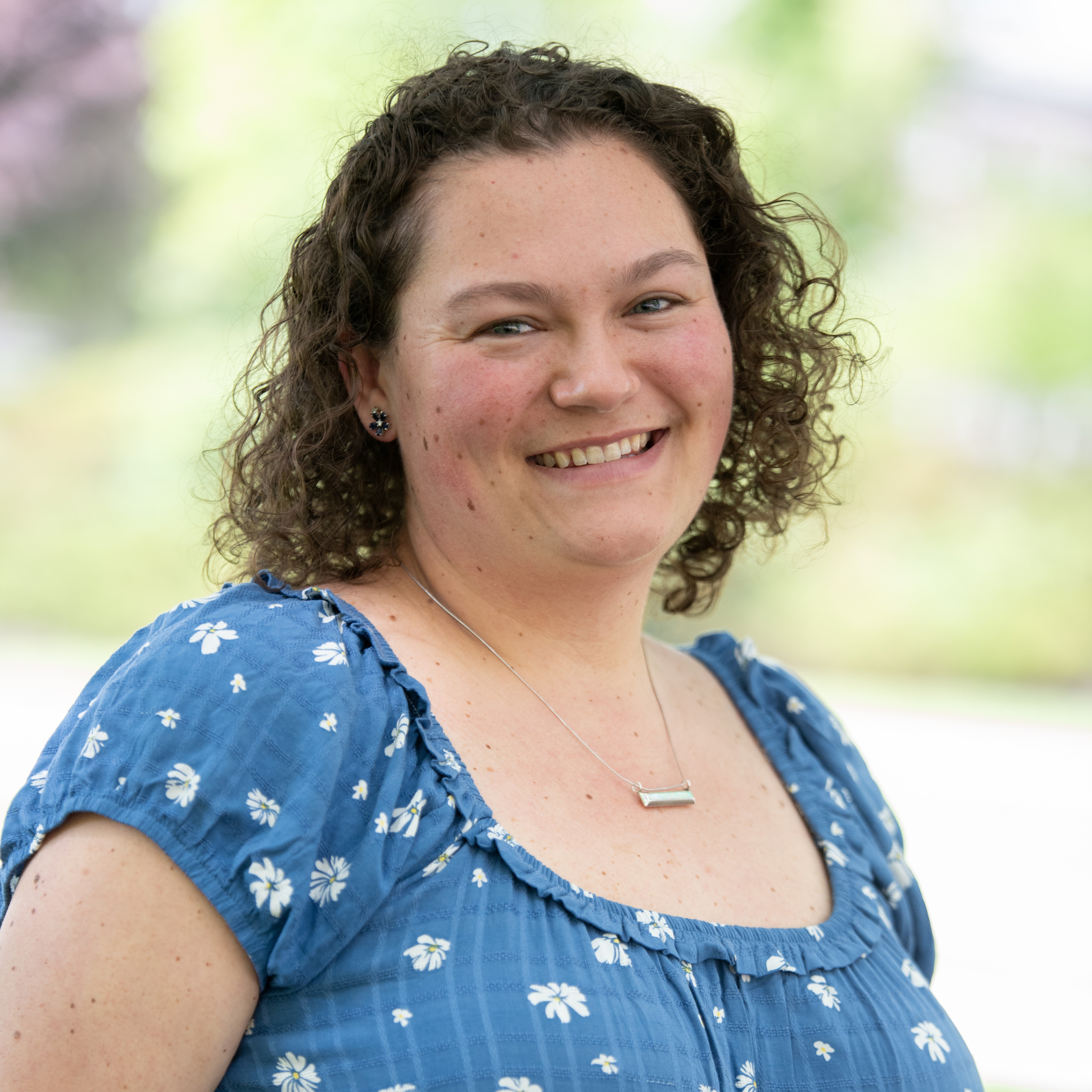 Close-up portrait of a person with short, curly hair, wearing a blue patterned top and a necklace, against a softly blurred outdoor background.