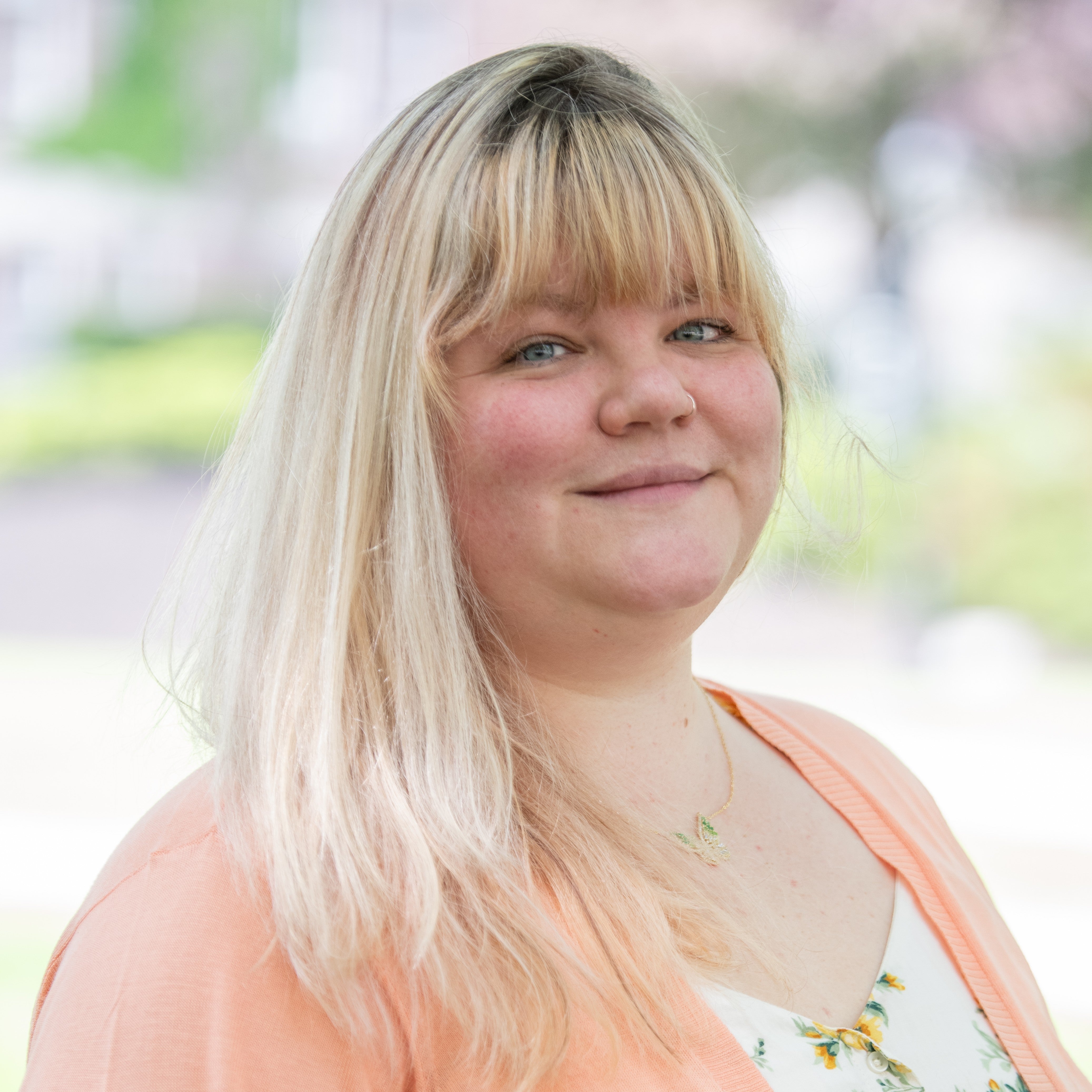 Close-up portrait of a person with long light hair and bangs, wearing a peach cardigan and a necklace, photographed outdoors with a softly blurred background.