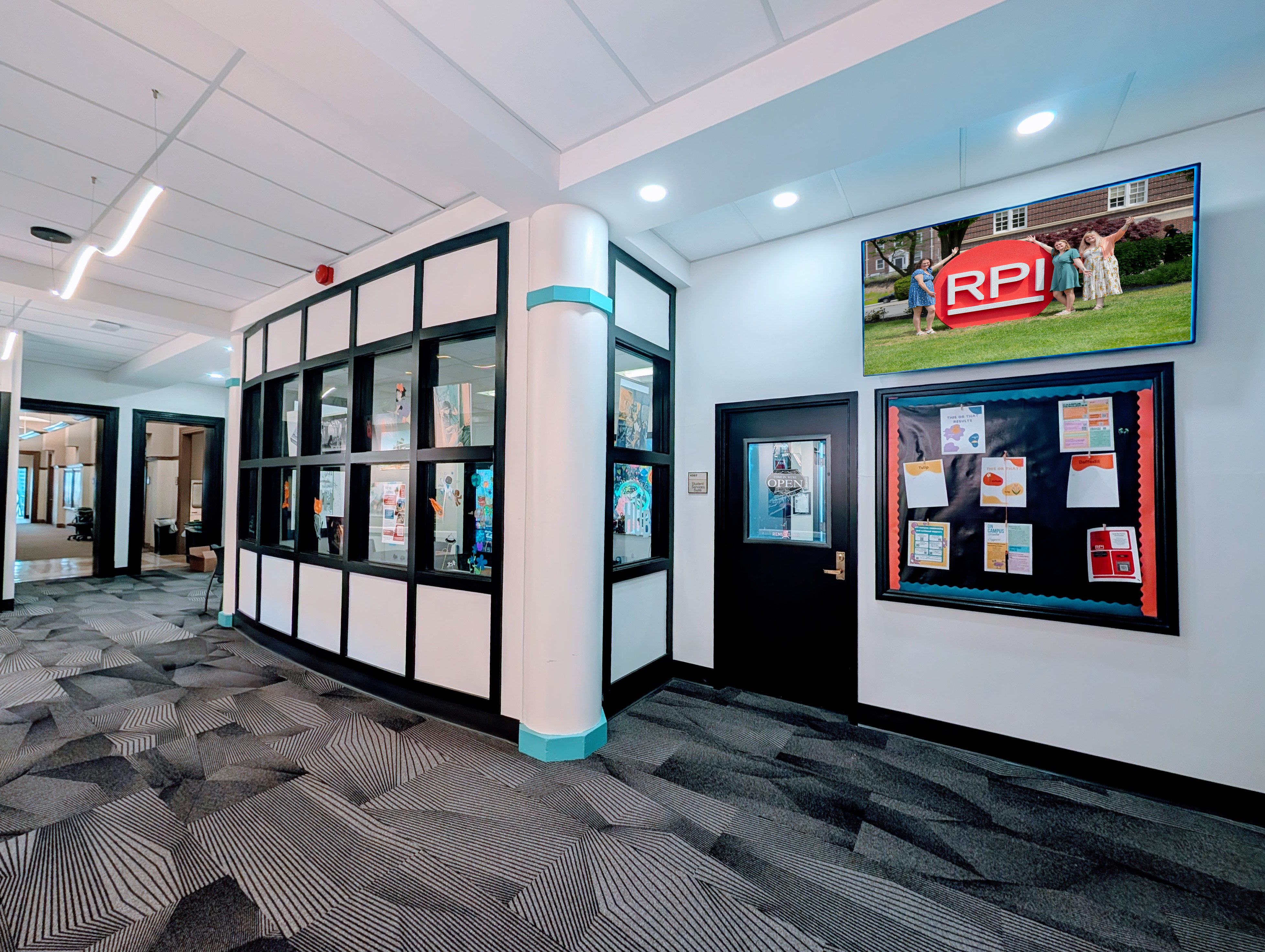 Modern indoor hallway with carpeted flooring, glass-walled offices, a closed door with an “OPEN” sign, a bulletin board with flyers, and a framed photo of people posing beside an RPI logo.