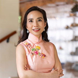 Catherine Tan, a dark haired woman wearing a pink dress, arms crossed