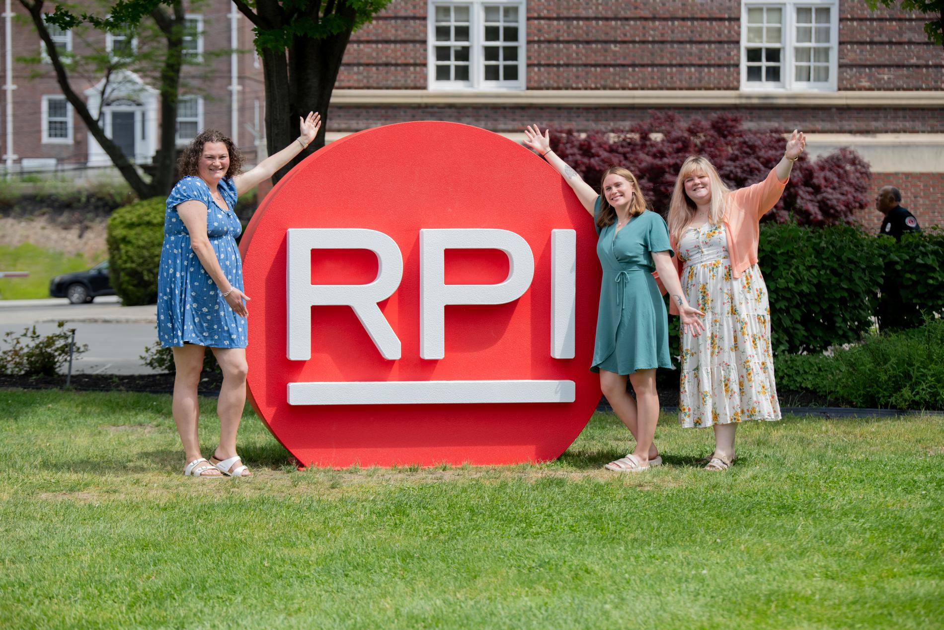 Three people stand outdoors on a lawn beside a large red circular sign with the white letters “RPI,” raising their arms while posing in front of a brick building and trees.