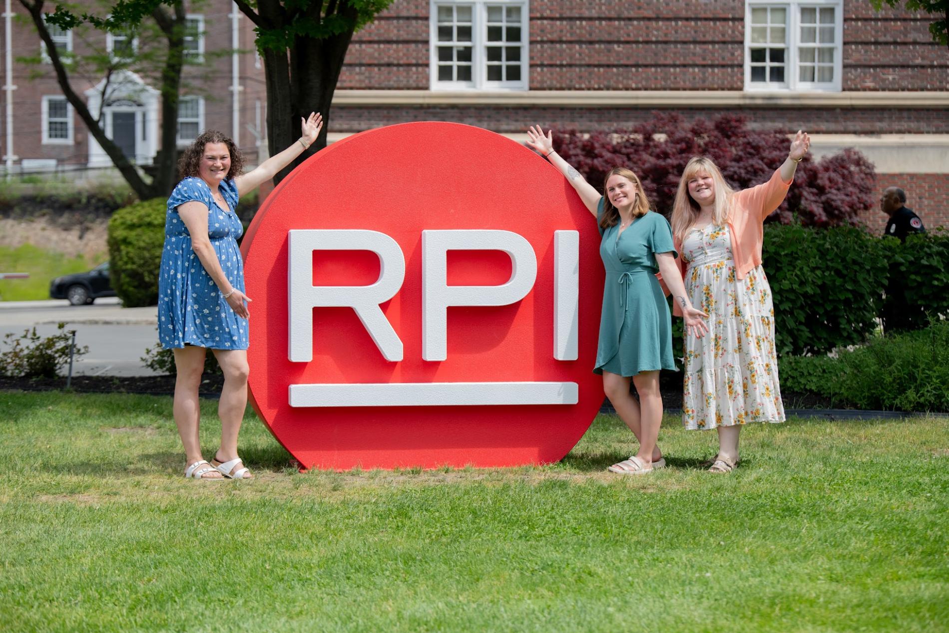 Three HASS Hub advisors stand around a large red RPI logo sign on a grassy area of campus, with brick academic buildings and trees in the background. The advisors are positioned on either side of the sign with their arms raised toward the logo.