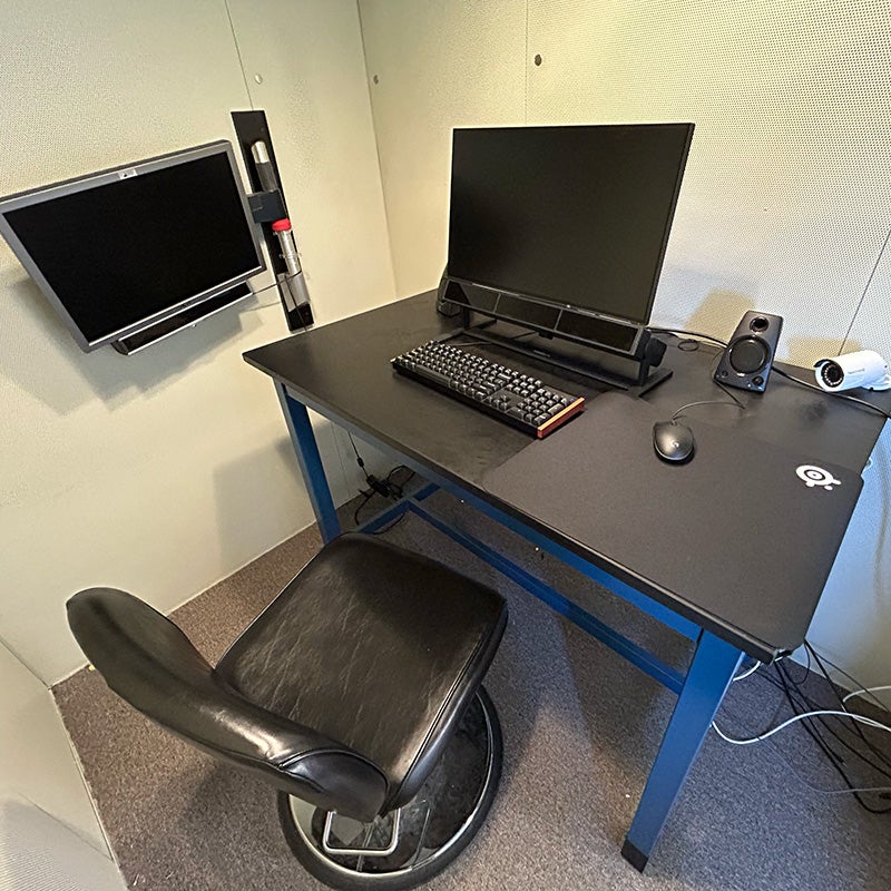 Computer workstation in a research lab with monitor, keyboard, mouse, speakers, and wall-mounted display inside a testing booth.