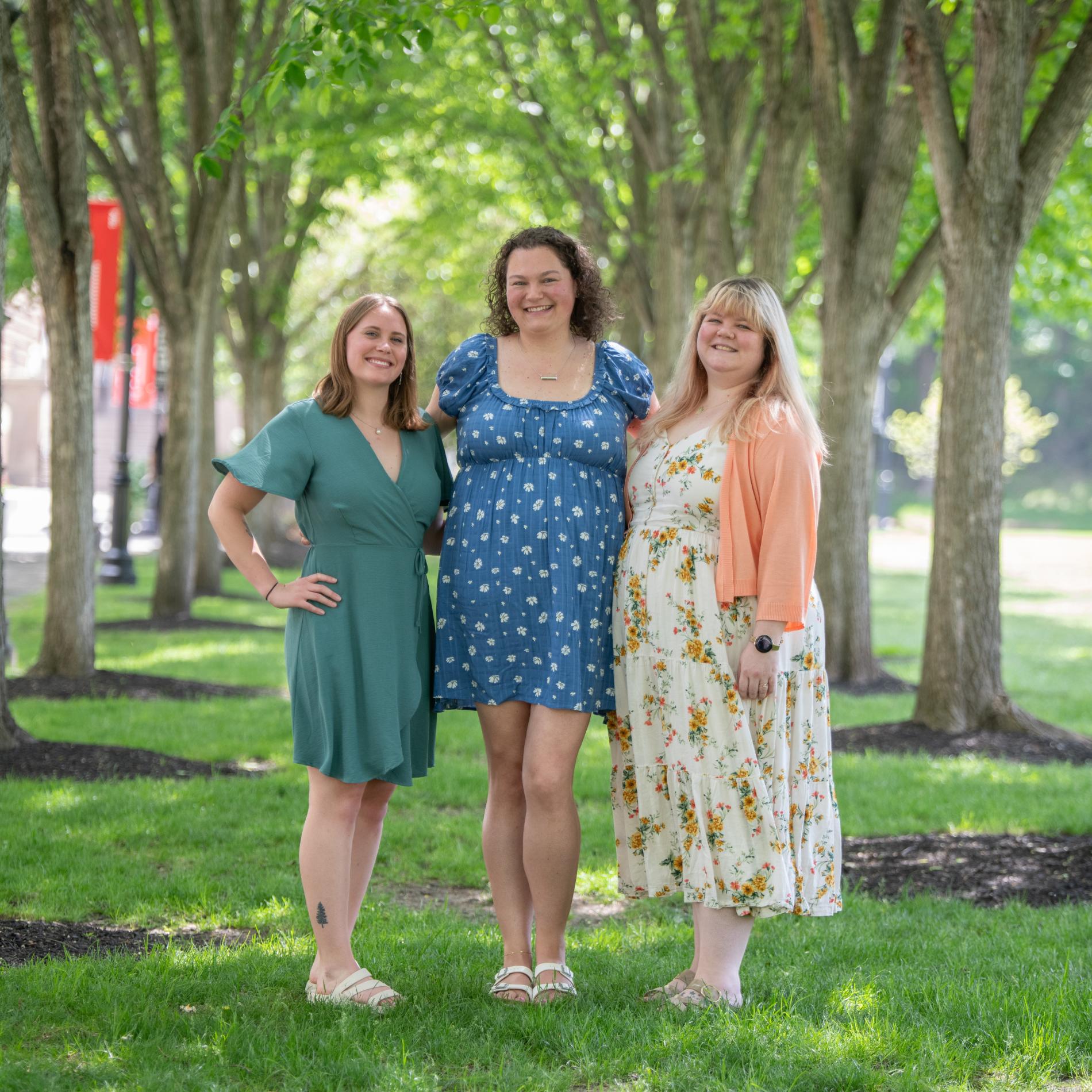 Three people stand side by side on a grassy lawn beneath a row of leafy trees, posing for a photo. Each person wears a casual dress—one green, one blue with a pattern, and one light-colored with a floral print and a light cardigan. Sunlight filters through the trees, and a campus-like walkway with a red banner is visible in the background