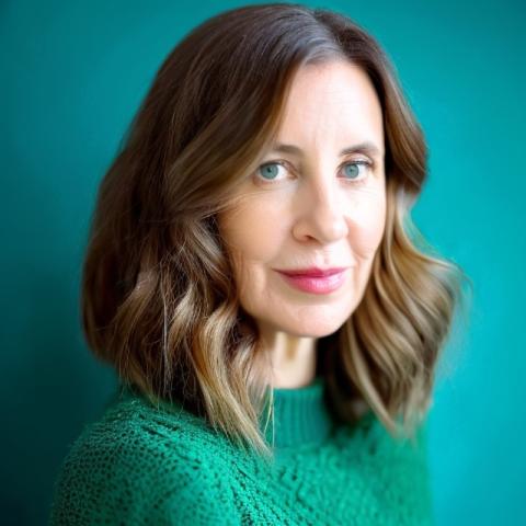 a caucasion woman with brown hair, wearing a green jumper, in front of a teal background