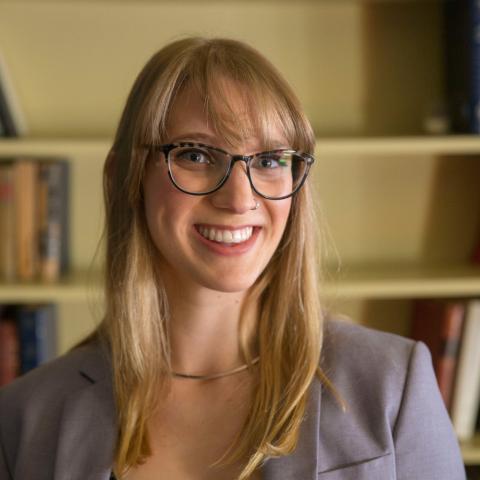 Portrait of a person with glasses smiling at the camera, standing indoors in front of a bookshelf filled with books.