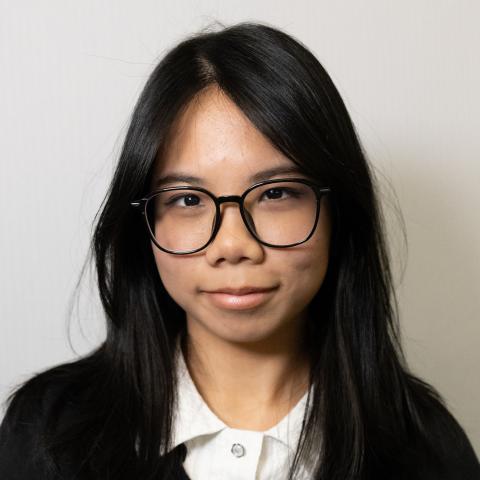 Close-up portrait of a person wearing round glasses and a collared shirt, looking at the camera against a plain, light background.