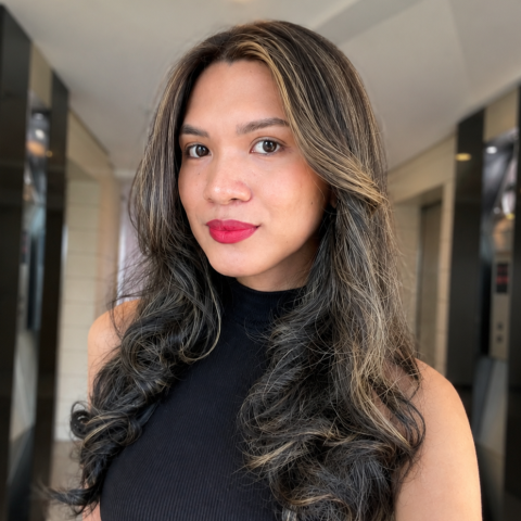 Close-up portrait of a person with long, dark wavy hair featuring light highlights, wearing a black sleeveless mock-neck top. The photo is taken indoors with a softly blurred hallway background.