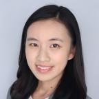 Professional headshot of a person with long dark hair, facing the camera against a light gray background, wearing a dark blazer over a light-colored top. The image is cropped from the shoulders up.