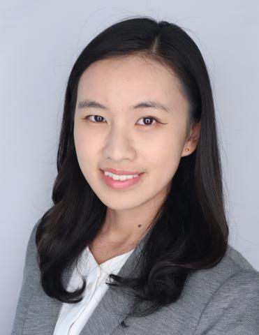 Professional headshot of a person with shoulder-length dark hair, wearing a gray blazer over a white collared blouse, facing the camera against a light gray studio background.