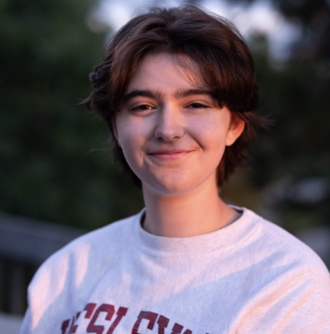 Close-up portrait of a student outdoors wearing a light-colored RPI sweatshirt, with trees softly blurred in the background.
