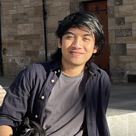 Person seated outdoors in bright sunlight, wearing a dark button-down shirt over a gray T-shirt. A stone building with a wooden door is visible in the background.