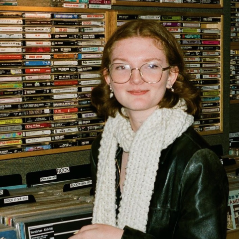 Person wearing round glasses, a white knitted scarf, and a black jacket standing in front of shelves filled with cassette tapes in a record store.