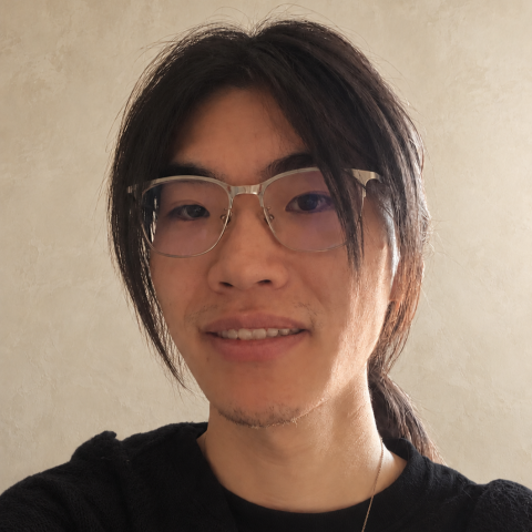 Portrait-style selfie of a person with long dark hair tied back, wearing clear-framed glasses, a black shirt, and a dark textured cardigan. The person is facing the camera indoors against a light beige wall. Soft natural light from the side illuminates the face and creates subtle shadows. A thin necklace chain is visible, and the image is closely cropped from the chest up. No text is visible in the image.